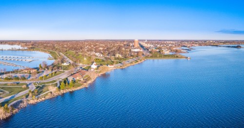 Aerial view of a serene waterfront with parks, buildings, and a clear blue sky reflecting on the water.
