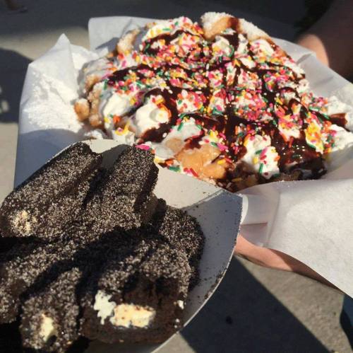 A hand holds a plate with a large dessert topped with whipped cream, sprinkles, and chocolate sauce, alongside chocolate treats.