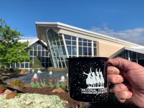 A hand holds a black mug with a cowboy design in front of the National Cowboy & Western Heritage Museum.