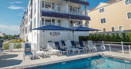 A pool area with lounge chairs and umbrellas in front of a white hotel building with balconies.