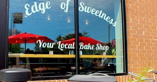 A bake shop window with "Edge of Sweetness" sign, outdoor seating, and red umbrellas in a sunny setting.