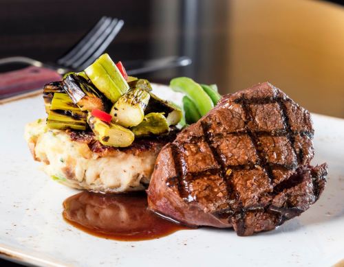 A plated dish featuring grilled steak, a vegetable-stuffed cake, and sautéed greens, garnished with sauce.