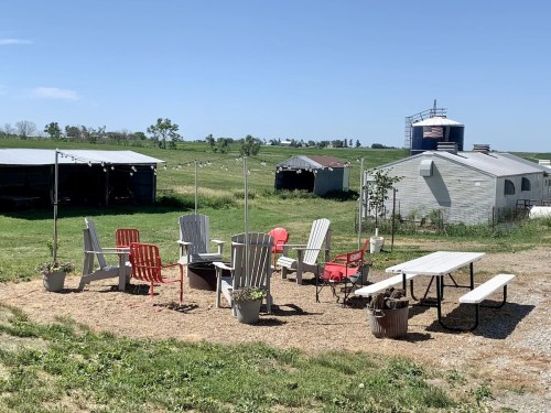 A cozy outdoor seating area with chairs and a table, surrounded by green fields and farm buildings under a clear blue sky.