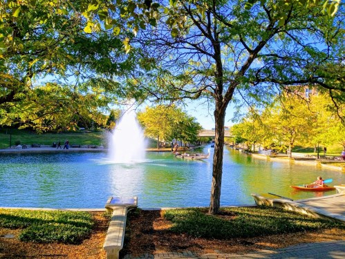 A serene park scene with a fountain in a pond, surrounded by trees and people enjoying the outdoors, including a kayaker.