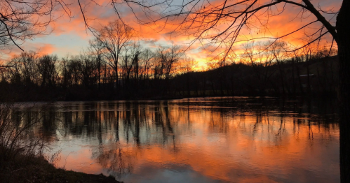 A serene river at sunset, reflecting vibrant orange and pink hues, framed by silhouetted trees.