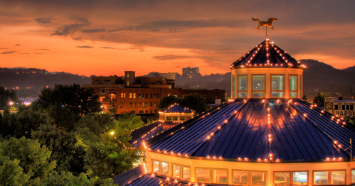 A sunset view of a carousel with a blue roof, adorned with lights, overlooking a cityscape and mountains in the background.