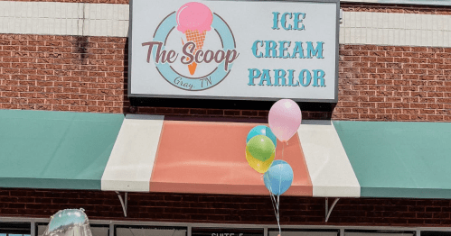 Sign for "The Scoop Ice Cream Parlor" with colorful balloons outside a brick building.
