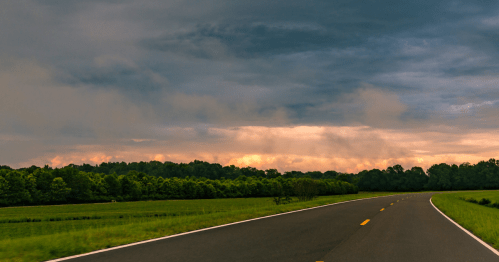 A winding road through lush green fields under a dramatic sky with dark clouds and a hint of sunset.