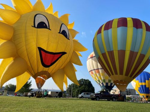 Colorful hot air balloons, including a large smiling sun, fill the sky against a clear blue backdrop.