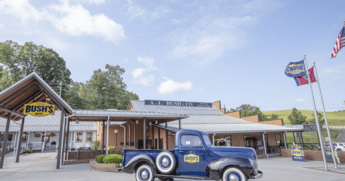 A vintage blue truck in front of the A.J. Bush Co. building, with flags and a clear sky in the background.