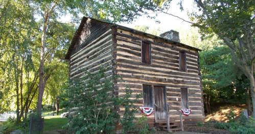 A rustic log cabin surrounded by trees, featuring a stone chimney and decorative bunting on the front.