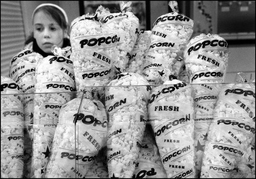 A girl looks at bags of fresh popcorn stacked in a display, captured in black and white.