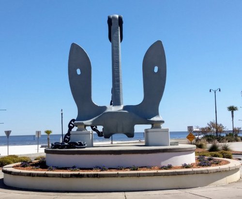 A large gray anchor sculpture stands on a circular flower bed by the waterfront under a clear blue sky.