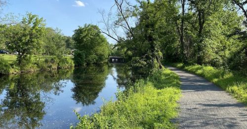 A serene pathway alongside a calm river, surrounded by lush greenery and trees under a clear blue sky.