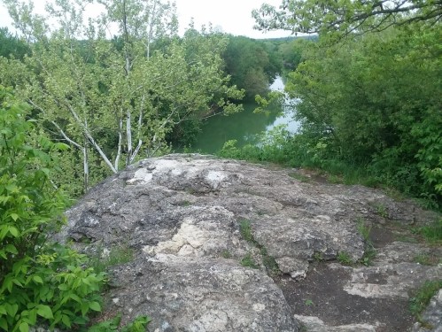 A rocky outcrop overlooks a lush green landscape and a calm river, surrounded by trees and foliage.