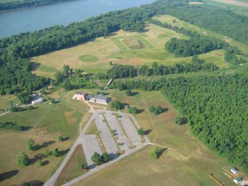Aerial view of a green landscape with fields, trees, and a river in the background, featuring a few buildings and parking areas.