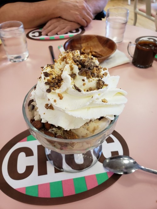 A glass dessert bowl filled with ice cream, whipped cream, and crumbled toppings, on a pink table with drinks and snacks.