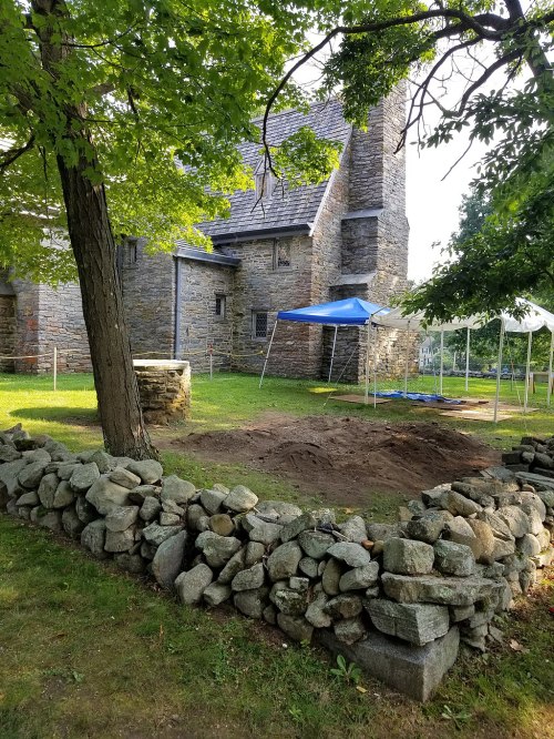 A stone wall surrounds a cleared area with tents, next to a historic stone building and trees in a grassy setting.