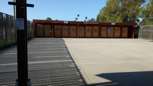 Abandoned building with boarded windows, surrounded by a fenced area and a clear blue sky. Shadows cast on the ground.