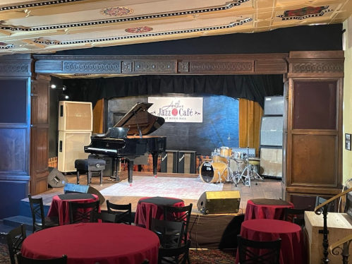 A stage set up for a performance, featuring a grand piano, drums, and tables with red tablecloths in a jazz café.