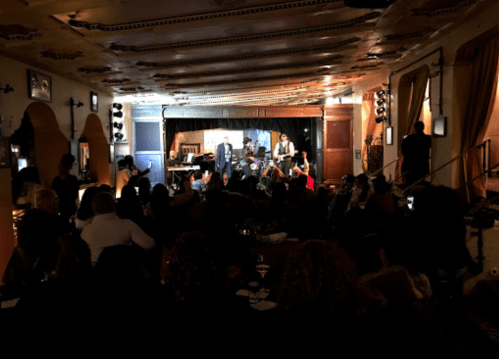 A lively performance on stage with musicians and singers, while an audience enjoys the show in a cozy venue.