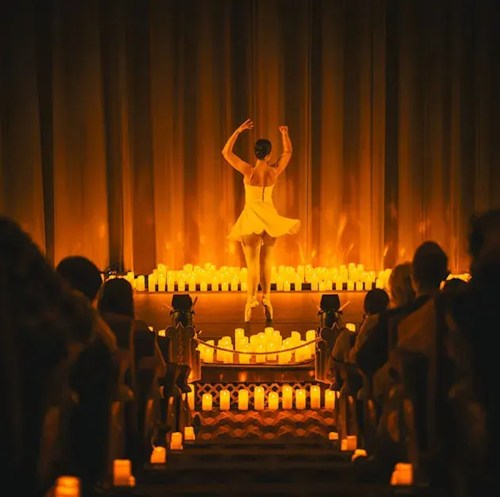 A dancer in a white dress performs on stage, surrounded by glowing candles and an audience in a dimly lit theater.