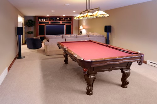 A spacious basement game room featuring a red felt pool table, a cozy sofa, and a TV area with decorative lighting.