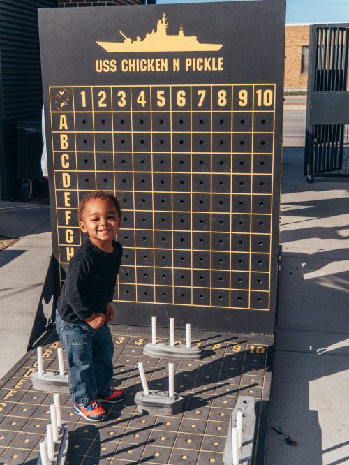 A smiling child stands next to a large game board labeled "USS Chicken N Pickle," ready to play.