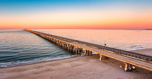 A long pier extends into calm waters at sunset, with a gradient sky of orange and blue hues.
