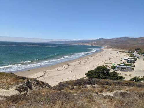 A scenic view of a sandy beach with gentle waves, surrounded by hills and a few coastal buildings in the distance.