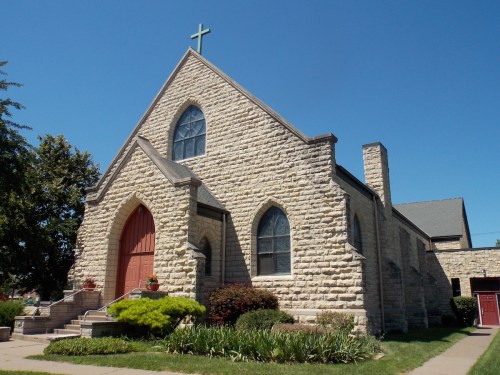 A stone church with a red door and cross on top, surrounded by greenery under a clear blue sky.
