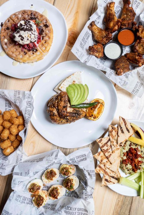A spread of delicious food including waffles, chicken wings, grilled chicken, tater tots, and pita bread on a wooden table.