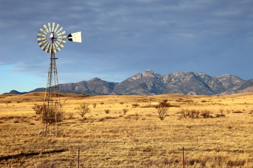 A windmill stands in a golden field with mountains in the background under a partly cloudy sky.