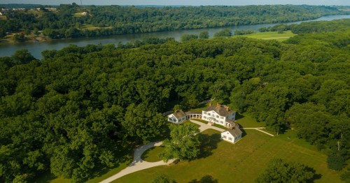 Aerial view of a large house surrounded by lush greenery and a river in the background.