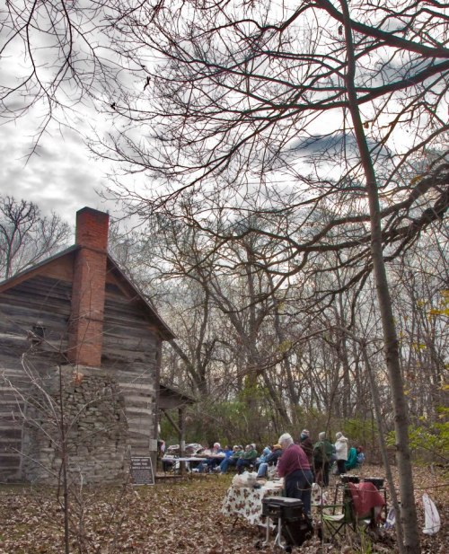 A group of people gathered outdoors near a rustic cabin, surrounded by bare trees and a cloudy sky.