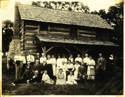 A large group of people poses in front of a rustic log cabin, dressed in early 20th-century clothing.