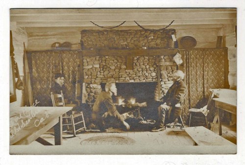 Three people seated in a rustic room with a stone fireplace, surrounded by wooden chairs and a cozy atmosphere.