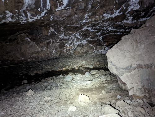 Dimly lit cave interior with rocky walls, a stream of water, and scattered stones on the ground.