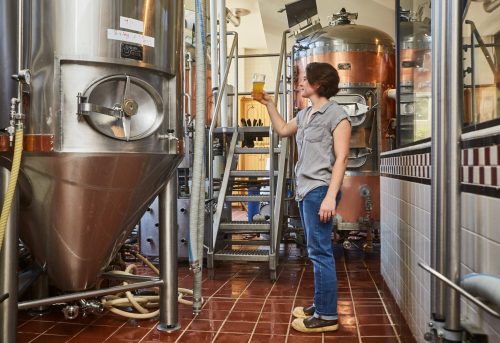 A woman holds a glass of beer in a brewery, surrounded by brewing equipment and stainless steel tanks.