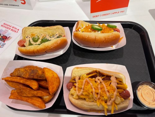 A tray with four hot dogs topped with various ingredients and a side of seasoned potato wedges.