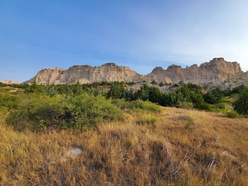 A scenic view of rugged cliffs rising above a grassy landscape with scattered trees under a clear blue sky.