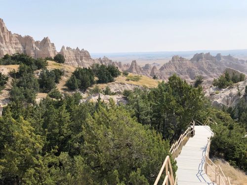 A wooden walkway winds through lush greenery, leading to rugged rock formations and a vast landscape in the distance.