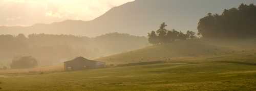 A serene landscape featuring a barn on a misty hillside, with mountains in the background and soft sunlight filtering through.