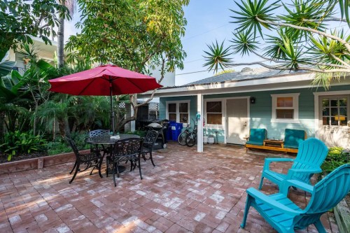 A sunny outdoor patio with a red umbrella, seating, and lush greenery surrounding a cozy blue house.