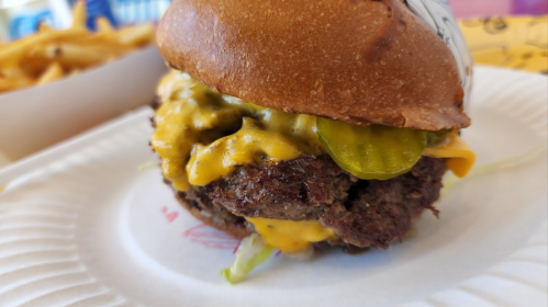 A close-up of a juicy cheeseburger with pickles and lettuce on a paper plate, with fries in the background.