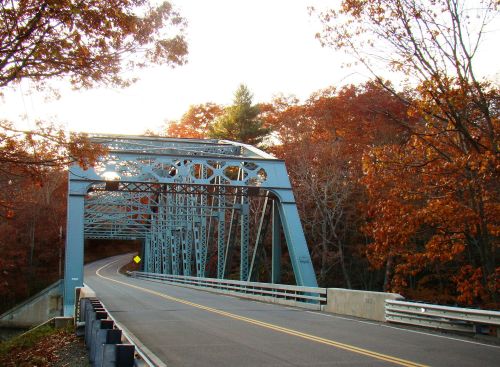 A blue metal bridge spans a winding road, surrounded by trees with autumn foliage in warm colors.