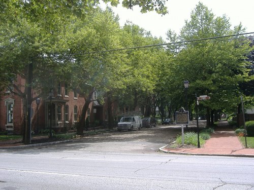 A tree-lined street with brick buildings, a parked van, and a historical marker on the corner.