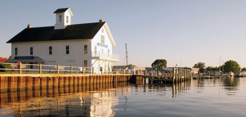 A white building with a steeple by a calm waterfront, reflecting in the water, with boats and a dock nearby.