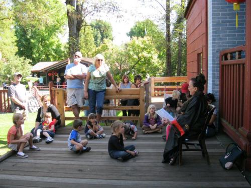 A storyteller in a black outfit reads to a group of children and adults seated on a wooden deck in a park.