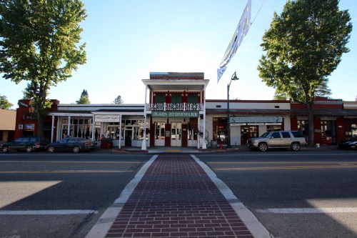 A charming street view of a small town with shops and trees lining the sidewalk under a clear blue sky.